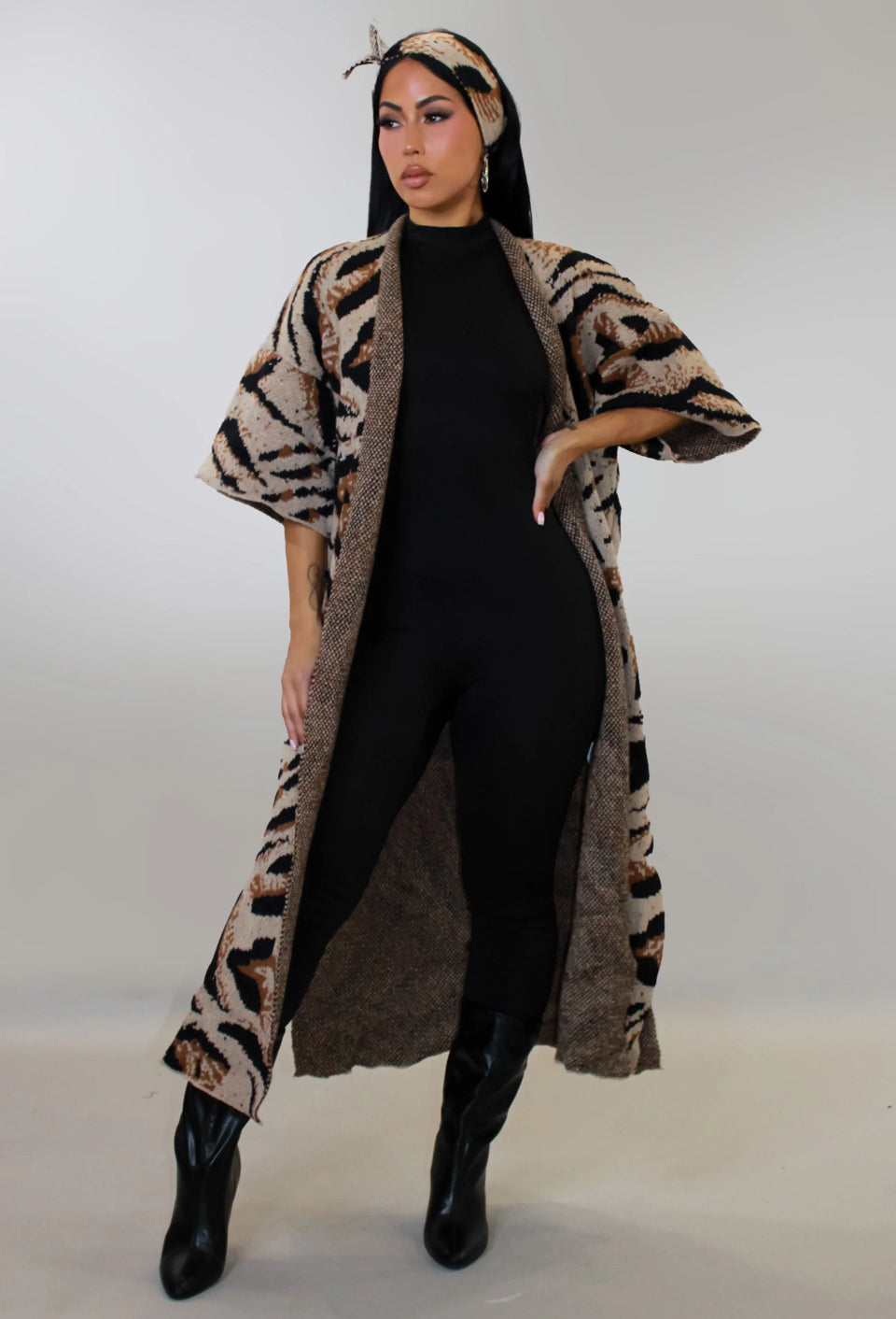 Woman wearing a long, patterned coat over a black outfit with black boots on a plain background